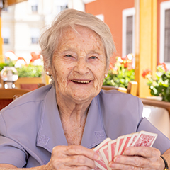 Natural Pharmaceuticals Hepactum FORTE Idős nő mosolyogva játszik kártyát napfényes teraszon, vidám és barátságos hangulatban.