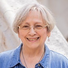 Natural Pharmaceuticals Hepactum FORTE Mosolygós idősebb nő, rövid ősz hajjal, szemüvegben, kék farmeringben, fehér háttér előtt.